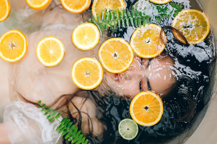 Person lying in water with sliced oranges and ferns around them